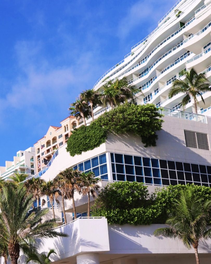 Modern beachfront building with palm trees and lush greenery against a bright blue sky, showcasing architectural design and coastal aesthetics.