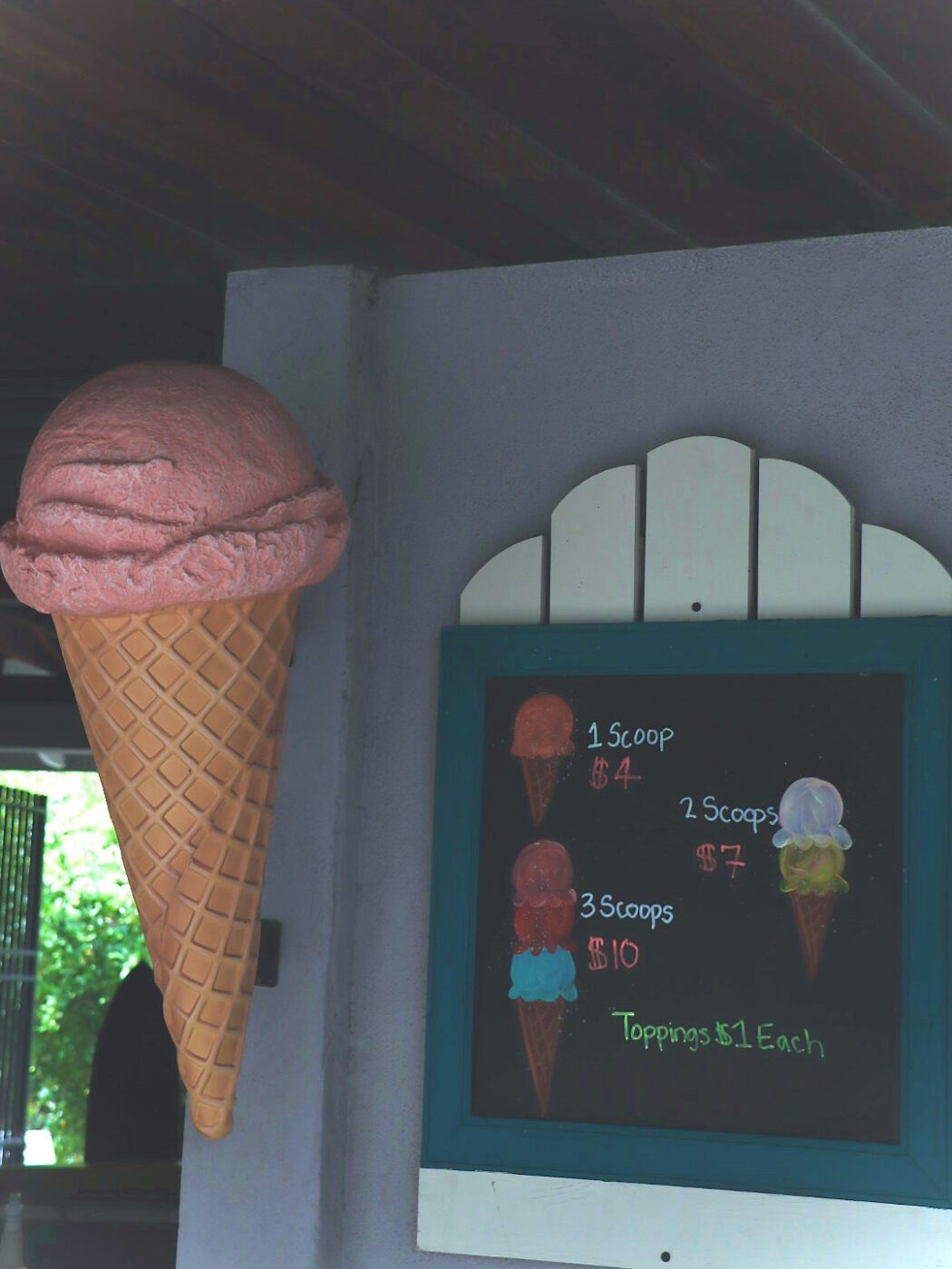Pink ice cream atop a large cone is displayed next to a chalkboard menu listing scoop prices: $4 for one, $7 for two, and $10 for three, with toppings priced at $1 each.
