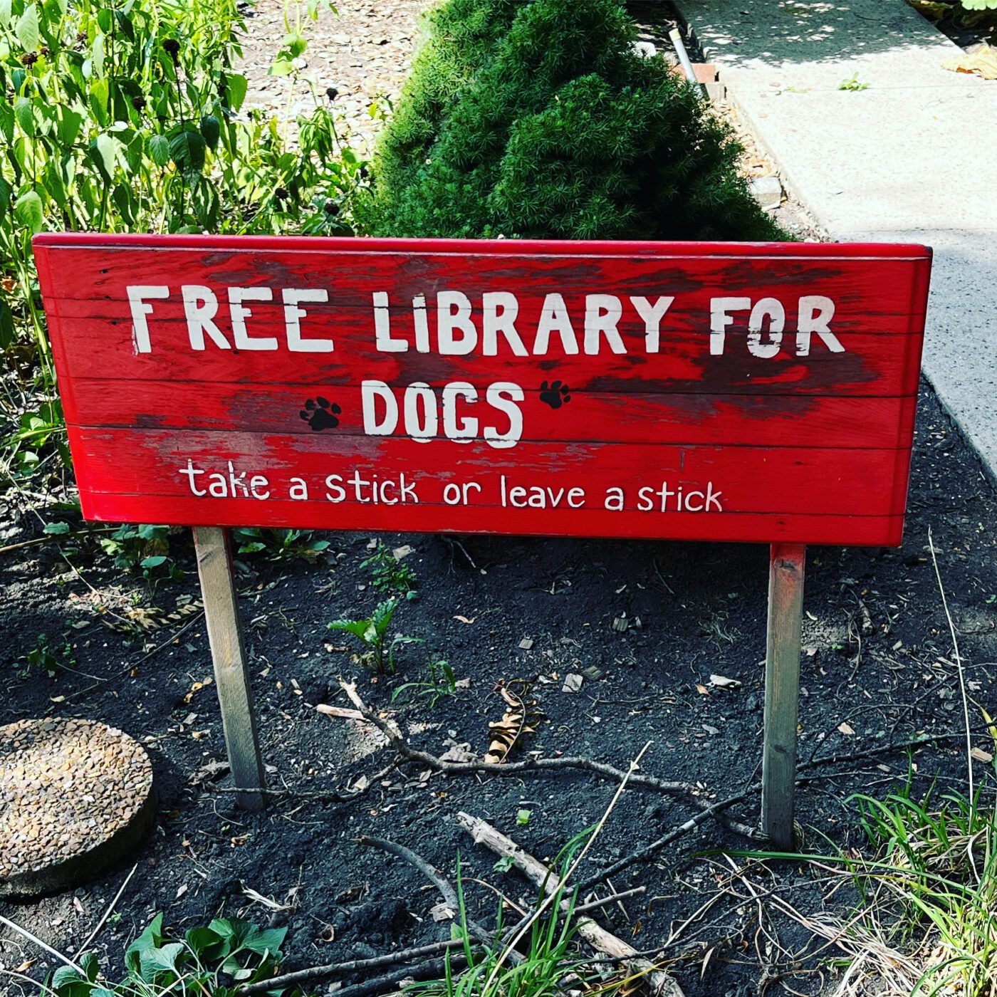 Red wooden sign reading "Free Library for Dogs" with the subtitle "take a stick or leave a stick," promoting playful interaction for dogs in a park.