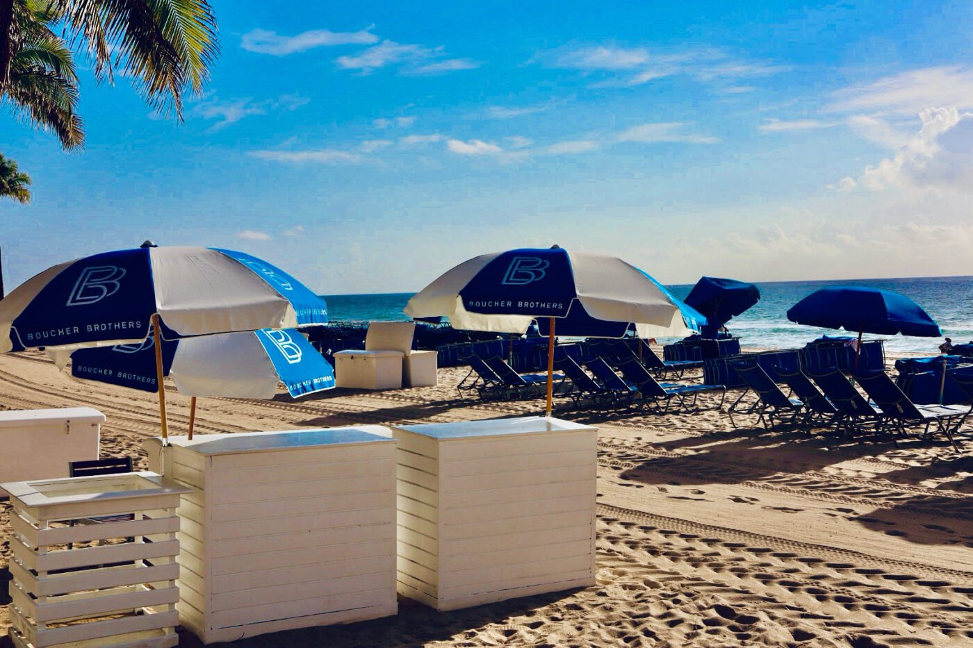 Blue-and-white umbrellas with "Boucher Brothers" shade rows of lounge chairs on a sunny beach, offering a relaxing seaside atmosphere.