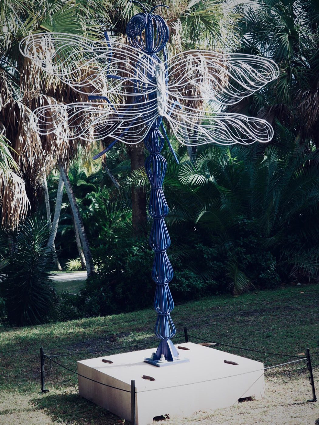 Large metal dragonfly sculpture stands amid tropical greenery, showcasing intricate wirework. It emphasizes nature and artistic expression in the park.