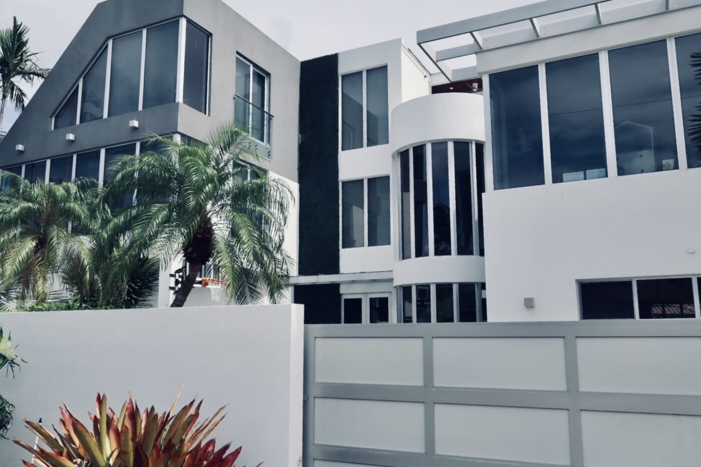 Modern architectural residence featuring large windows, palm trees, and a basketball hoop, set behind a white fence in an upscale area.