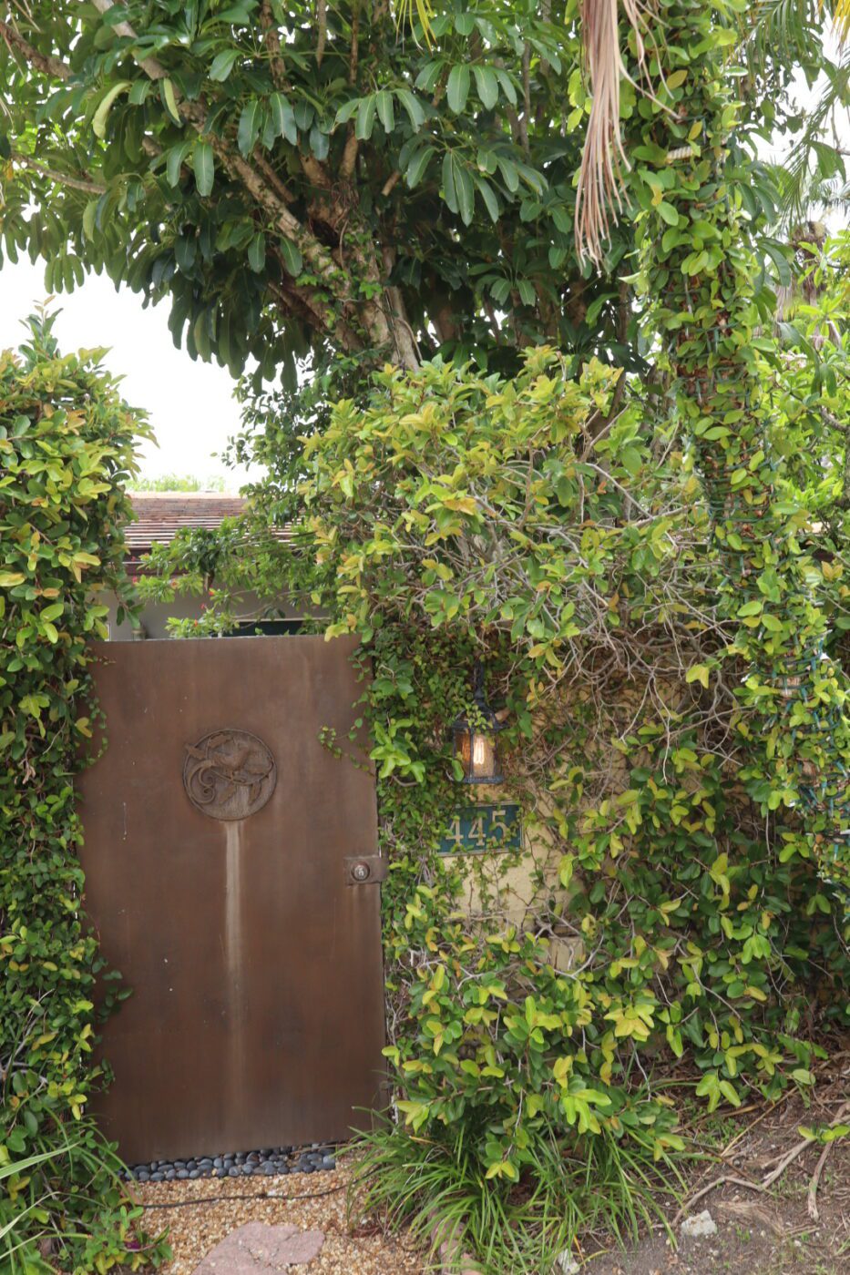 Rustic metal gate surrounded by lush greenery leads to a hidden entrance, with a visible house number (145) and an outdoor lantern.