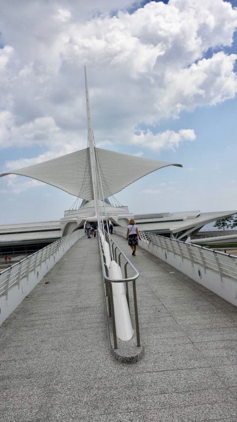 Architectural bridge with a sail-like design, connecting to the Milwaukee Art Museum. Visitors walk along the pathway under a blue sky.