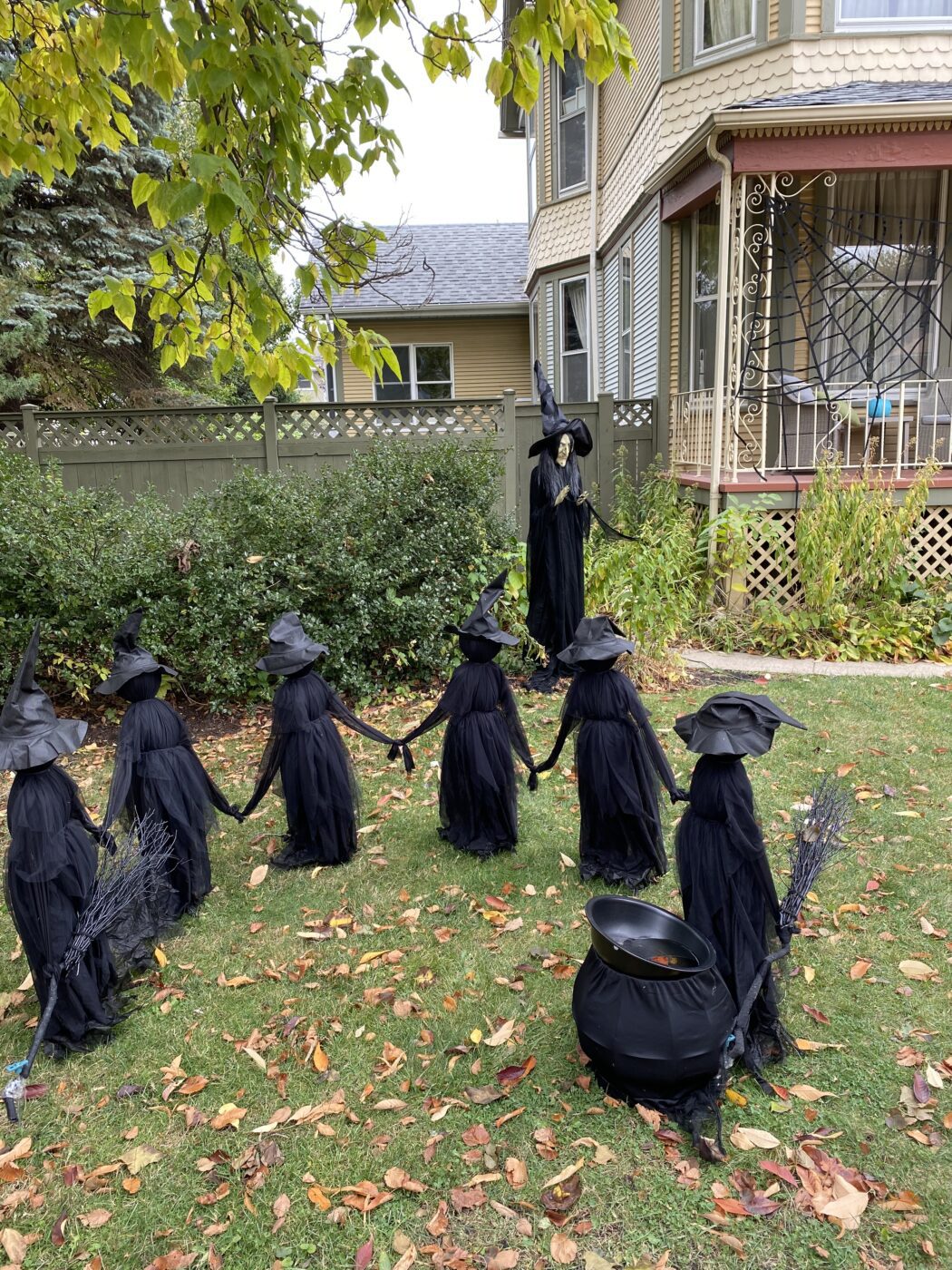 A group of six witch decorations in black cloaks and hats stand hand-in-hand in a yard, with a life-sized witch figure and cauldron nearby.