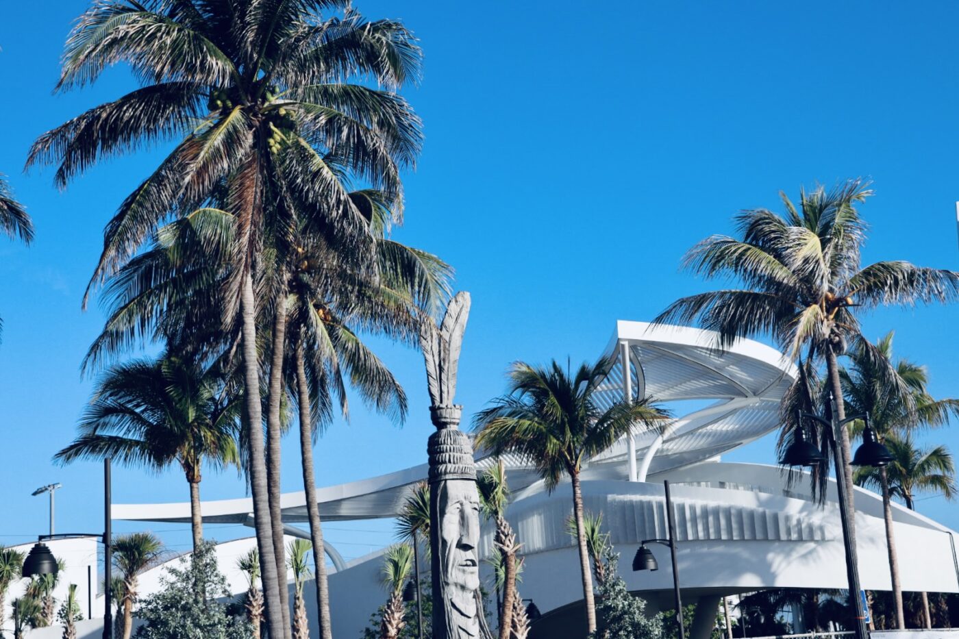 Palm trees frame a towering sculpture resembling a Native American figure in front of a contemporary, curving building against a clear blue sky.
