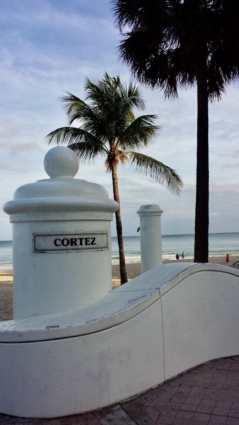 Cortez beach entrance features white decorative pillars, palm trees, and a view of the ocean, inviting visitors to enjoy the shoreline.