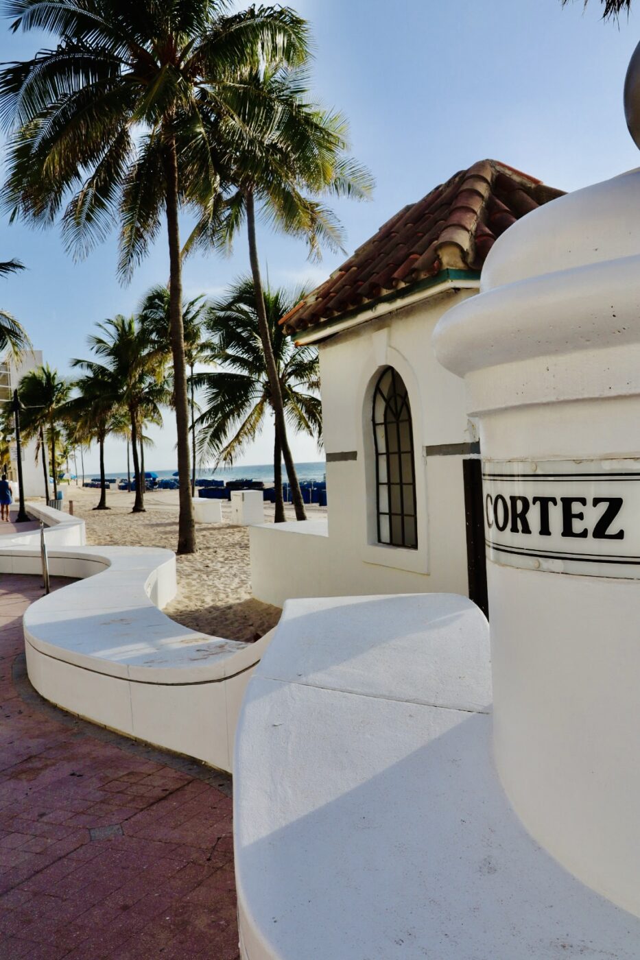 Seaside path lined with palm trees, leading to a sandy beach. A white structure labeled "Cortez" is visible, emphasizing the coastal atmosphere.