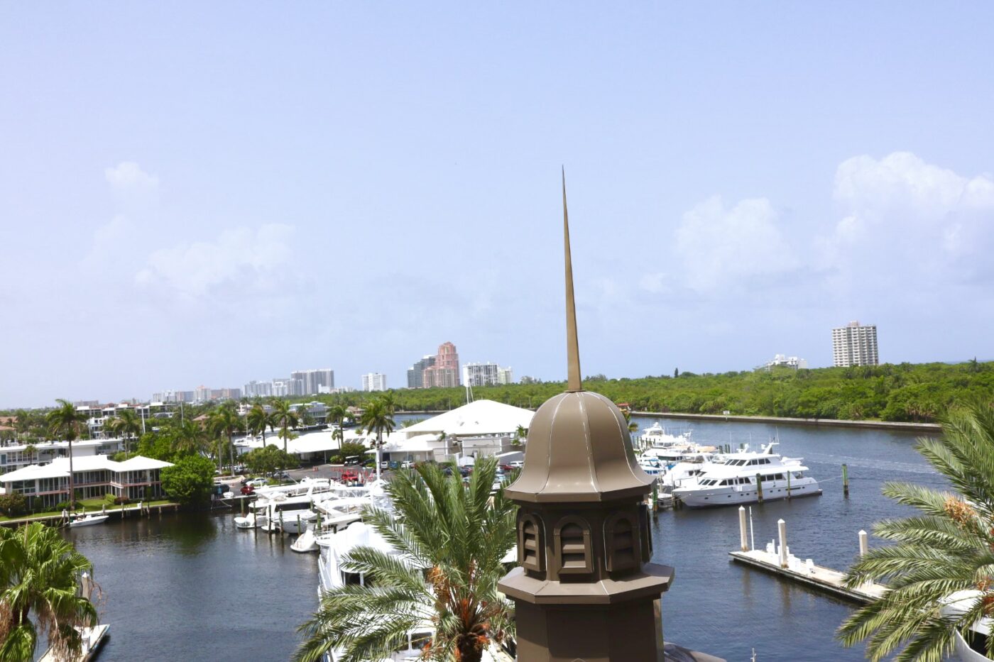 Scenic view featuring a marina with white yachts, lush greenery, and city skyline in the distance, framed by a decorative rooftop spire.