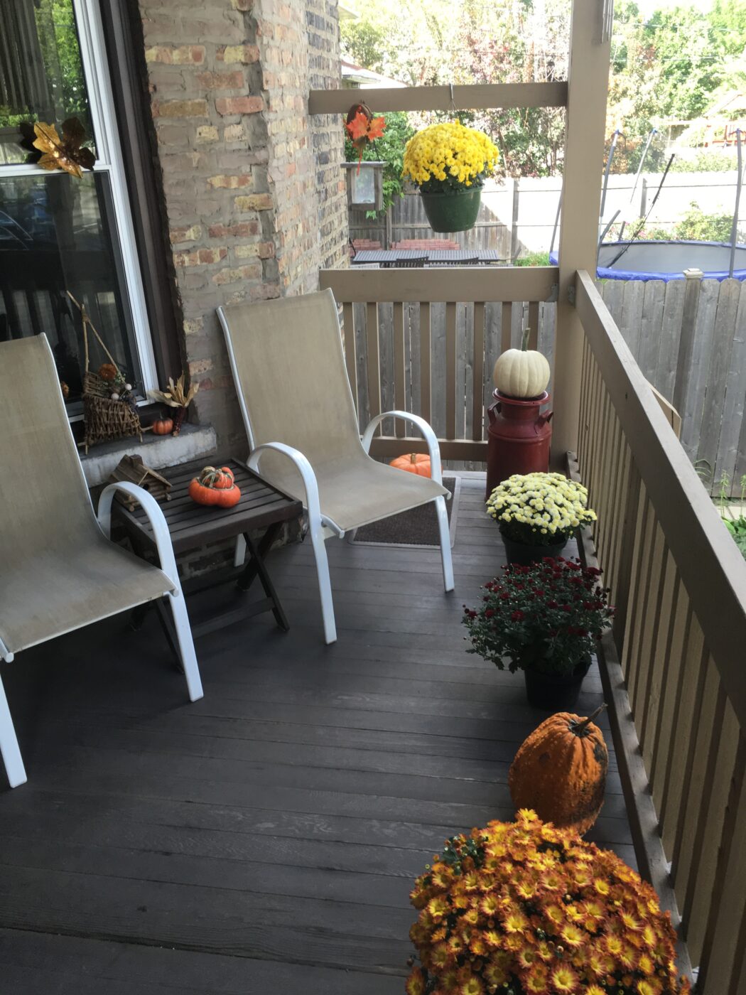 A cozy porch features two chairs, a small table, and decorative autumn flowers and pumpkins, creating a warm fall atmosphere.