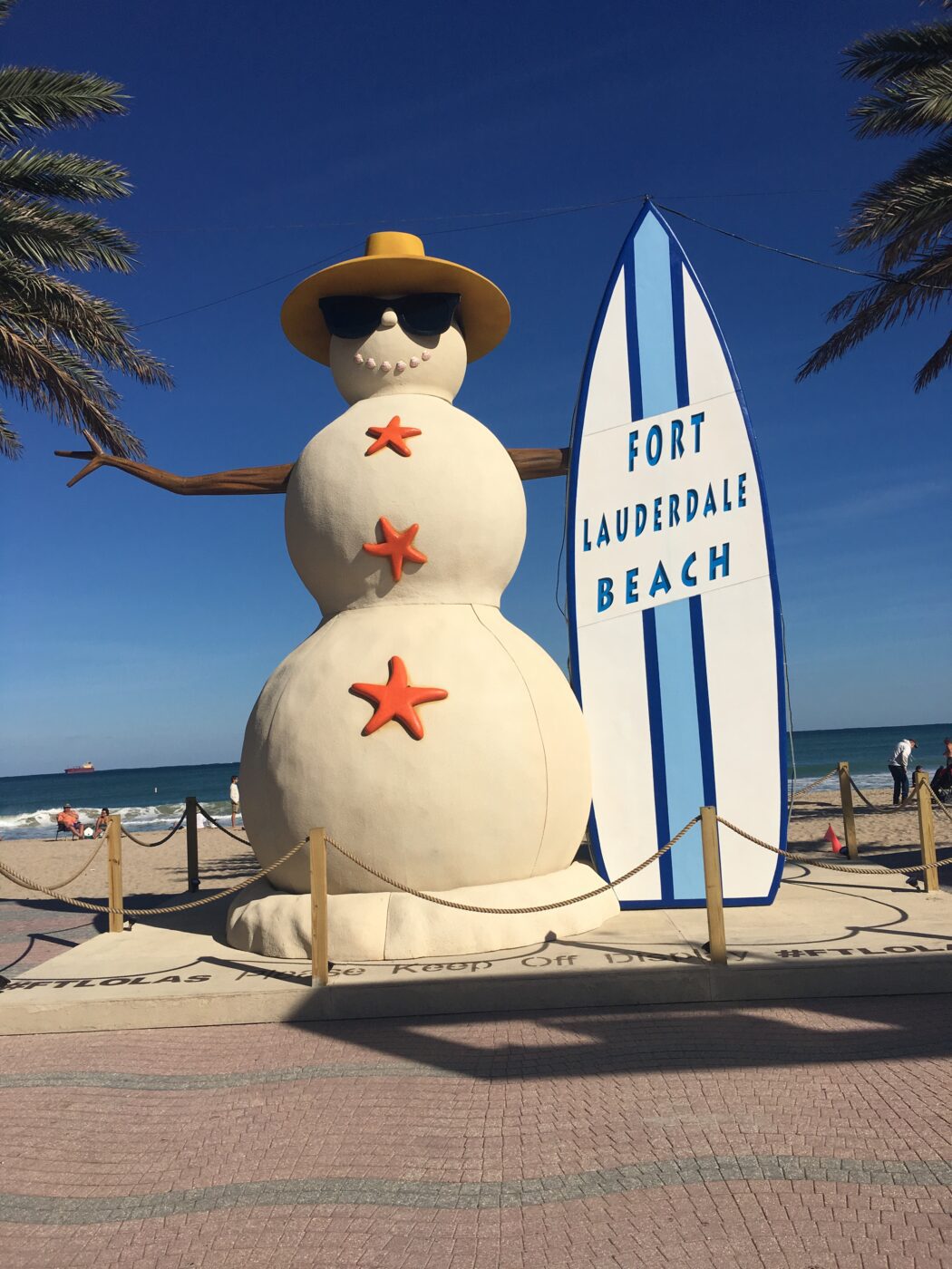 A large snowman in sunglasses and a hat stands beside a surfboard sign for Fort Lauderdale Beach, set against a clear blue sky.