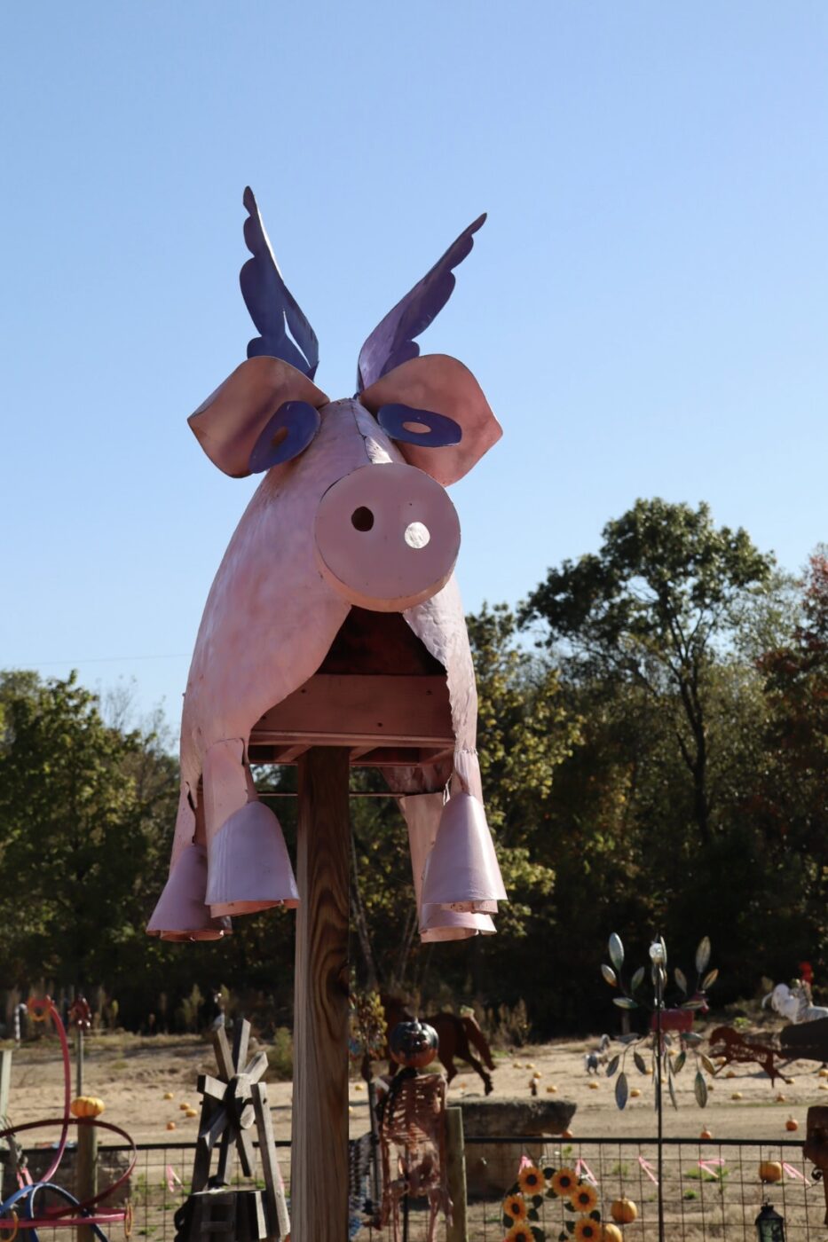 A whimsical metal pig sculpture with wings stands atop a post, surrounded by a colorful garden art display, highlighting creativity and fun in outdoor decor.