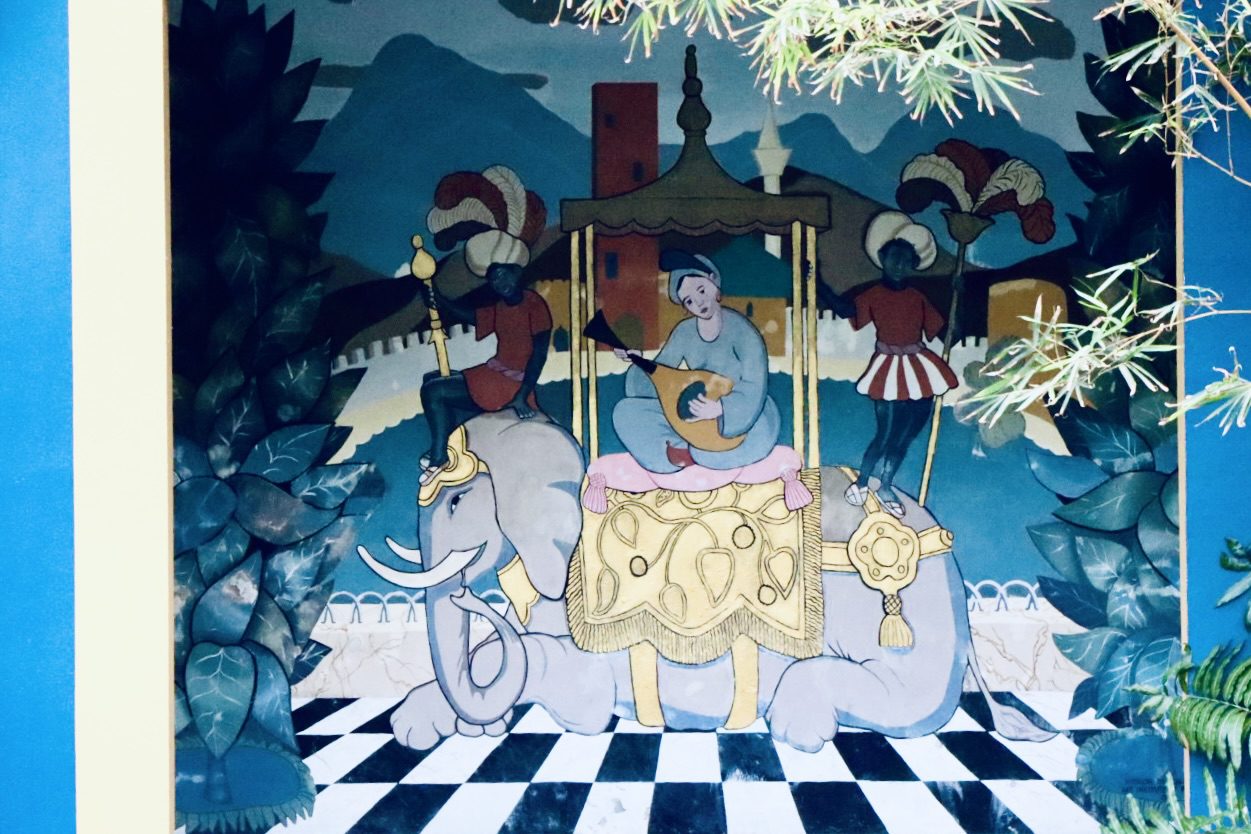 A colorful mural featuring a seated musician playing a lute on an adorned elephant, accompanied by two attendants. The backdrop includes mountains and architectural elements, capturing a whimsical scene.