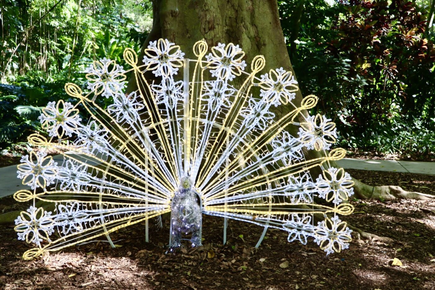A decorative peacock sculpture made of white and yellow lights, set against a lush, green background of trees and foliage.