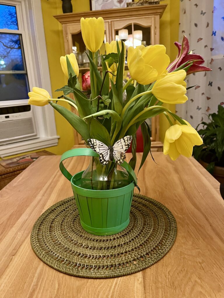 Bright yellow tulips and pink lilies in a green bucket-style vase. The arrangement is set on a woven placemat against a warm yellow wall.