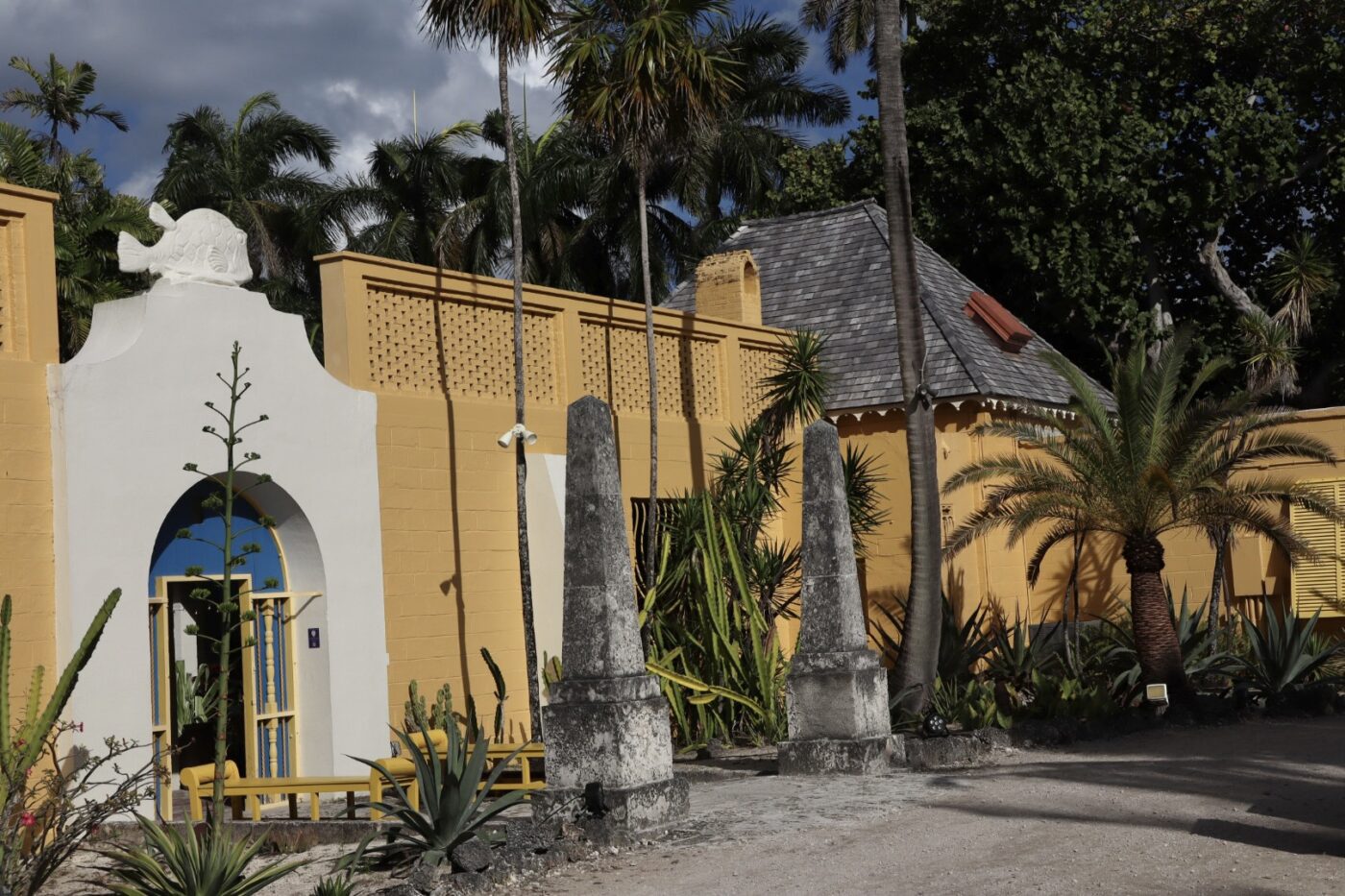 Colorful entrance featuring a white archway and yellow walls, adorned with tropical plants and stone pillars, representing Caribbean architecture.