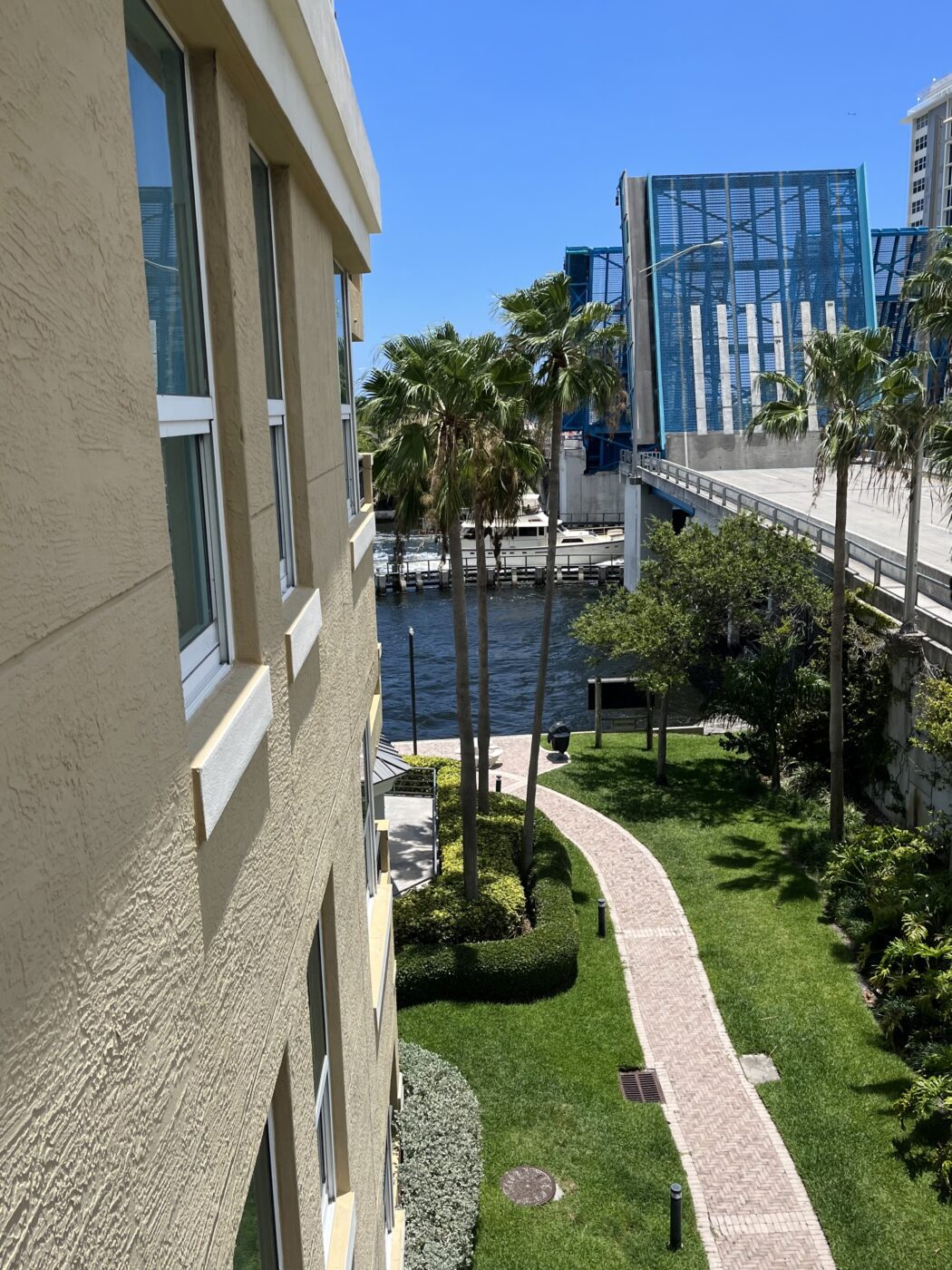 View from a building overlooking a pathway with palm trees, leading to a canal and a raised bridge against a clear blue sky.