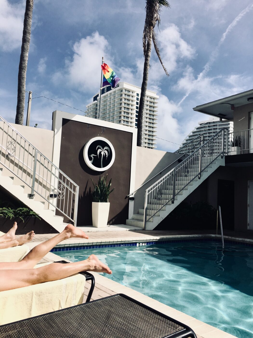 Sunny poolside scene featuring relaxation chairs, a rainbow flag waving, and modern buildings in the background, reflecting an inviting atmosphere.