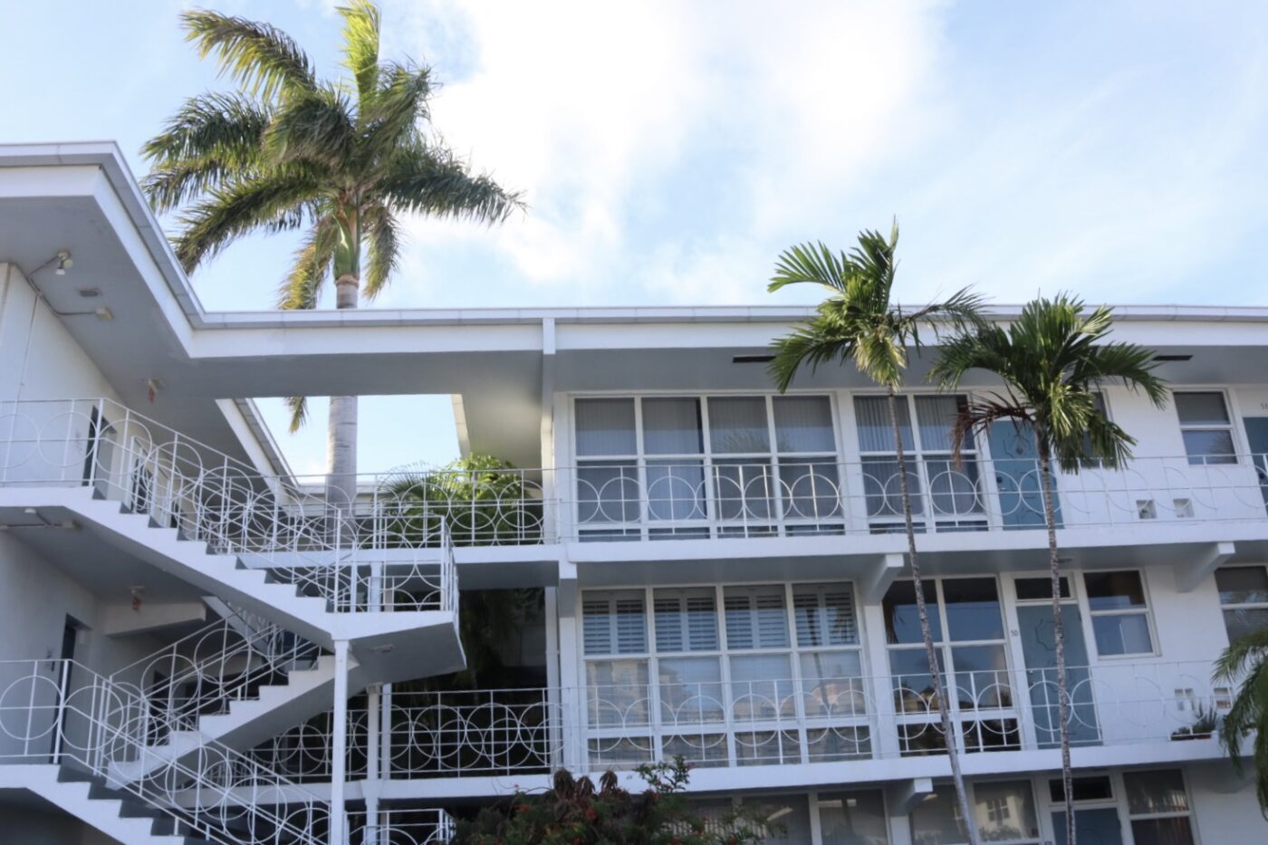 Modern hotel building with white balconies and spiral staircases, surrounded by palm trees under a blue sky, suggesting a tropical getaway.