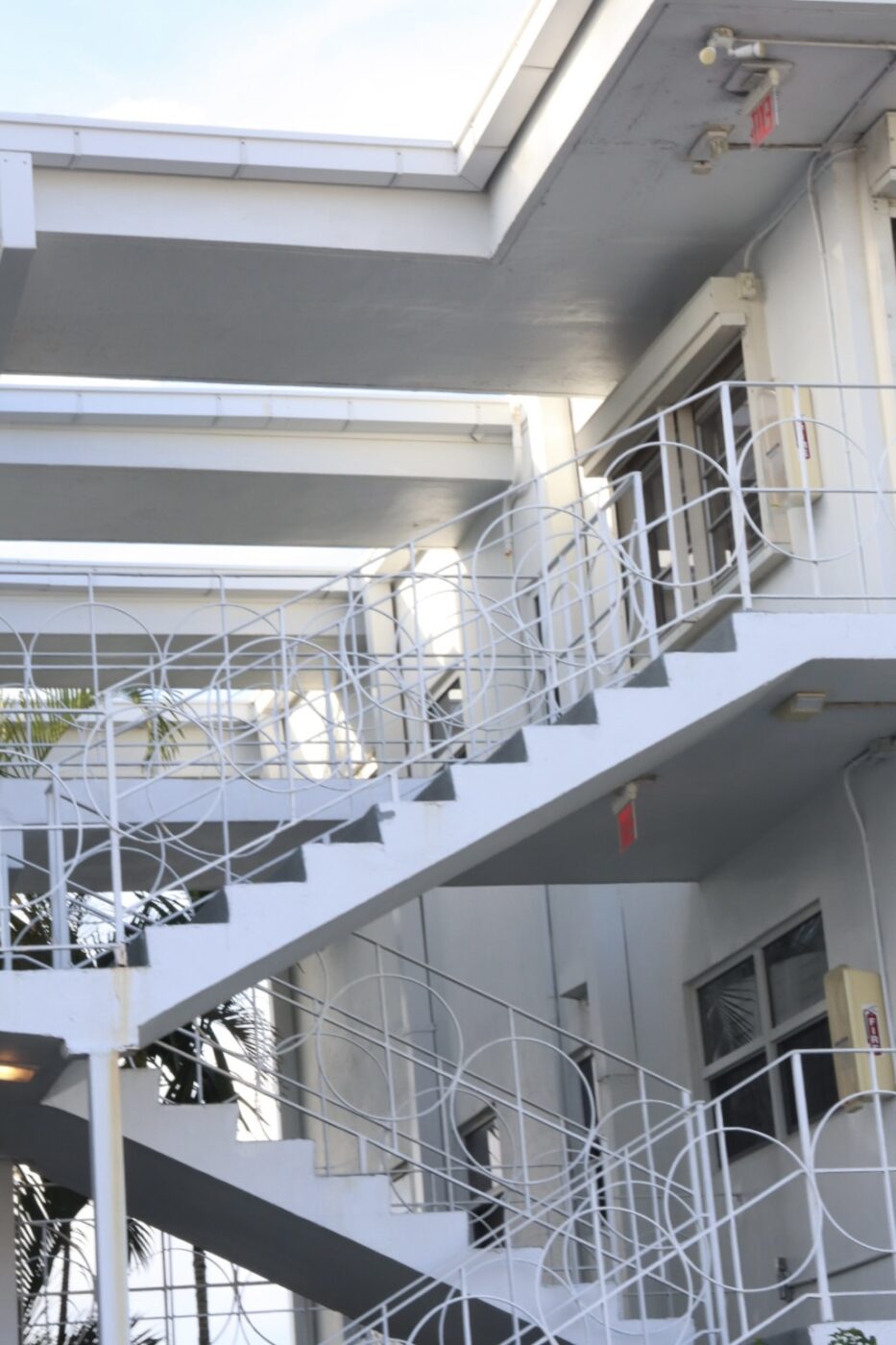 White exterior stairway with decorative railing, leading to upper-level doors in a multi-story building surrounded by palm trees.