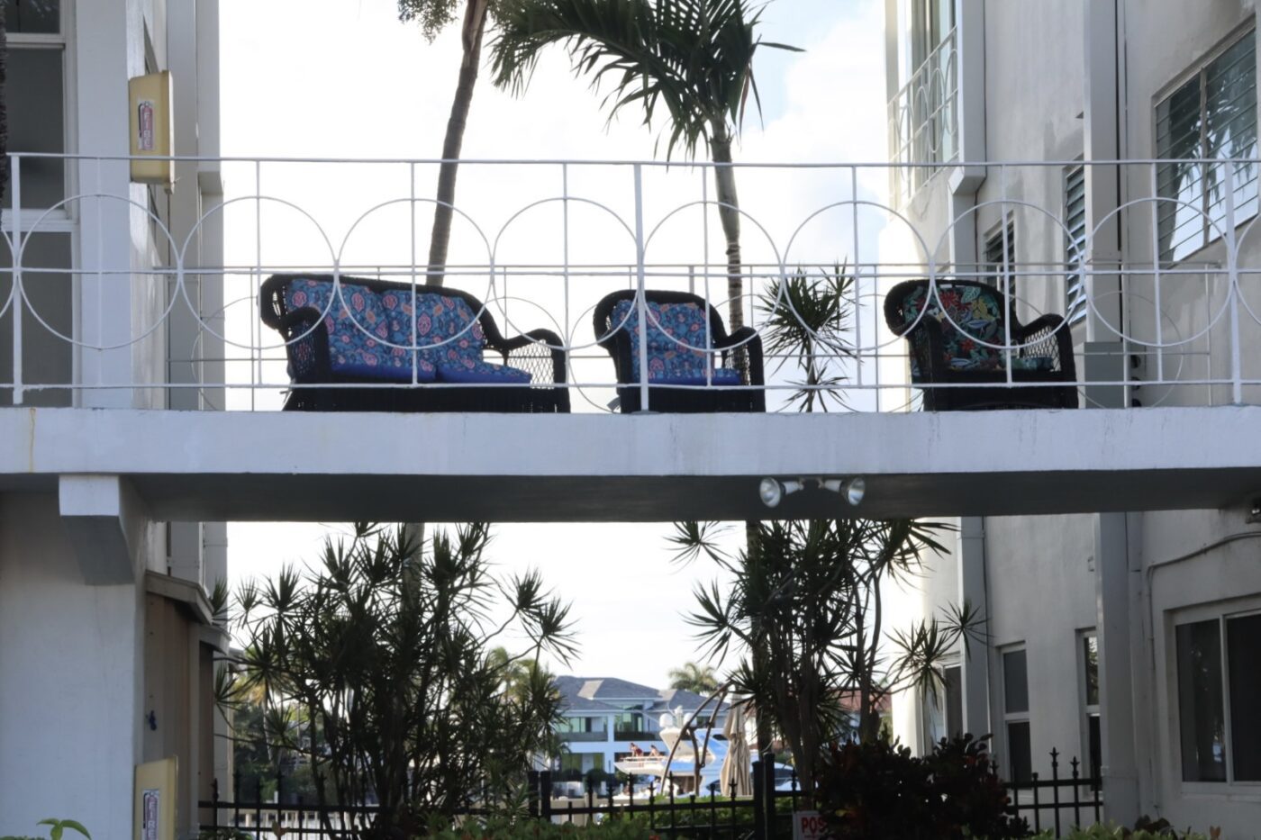 Colorful couches with floral patterns line a white railing on a bridge overlooking palm trees and a marina, reflecting a coastal vibe.