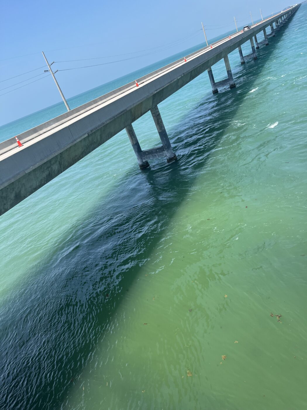A long bridge extends over vibrant turquoise waters, with concrete supports casting shadows below. Traffic cones indicate ongoing work along the structure.