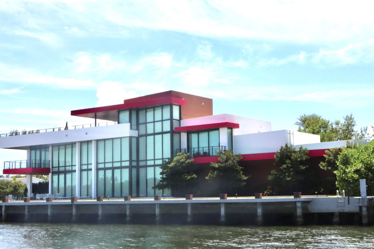 Modern waterfront building featuring large glass windows and a striking red roof. Surrounded by greenery, this structure highlights contemporary architecture.