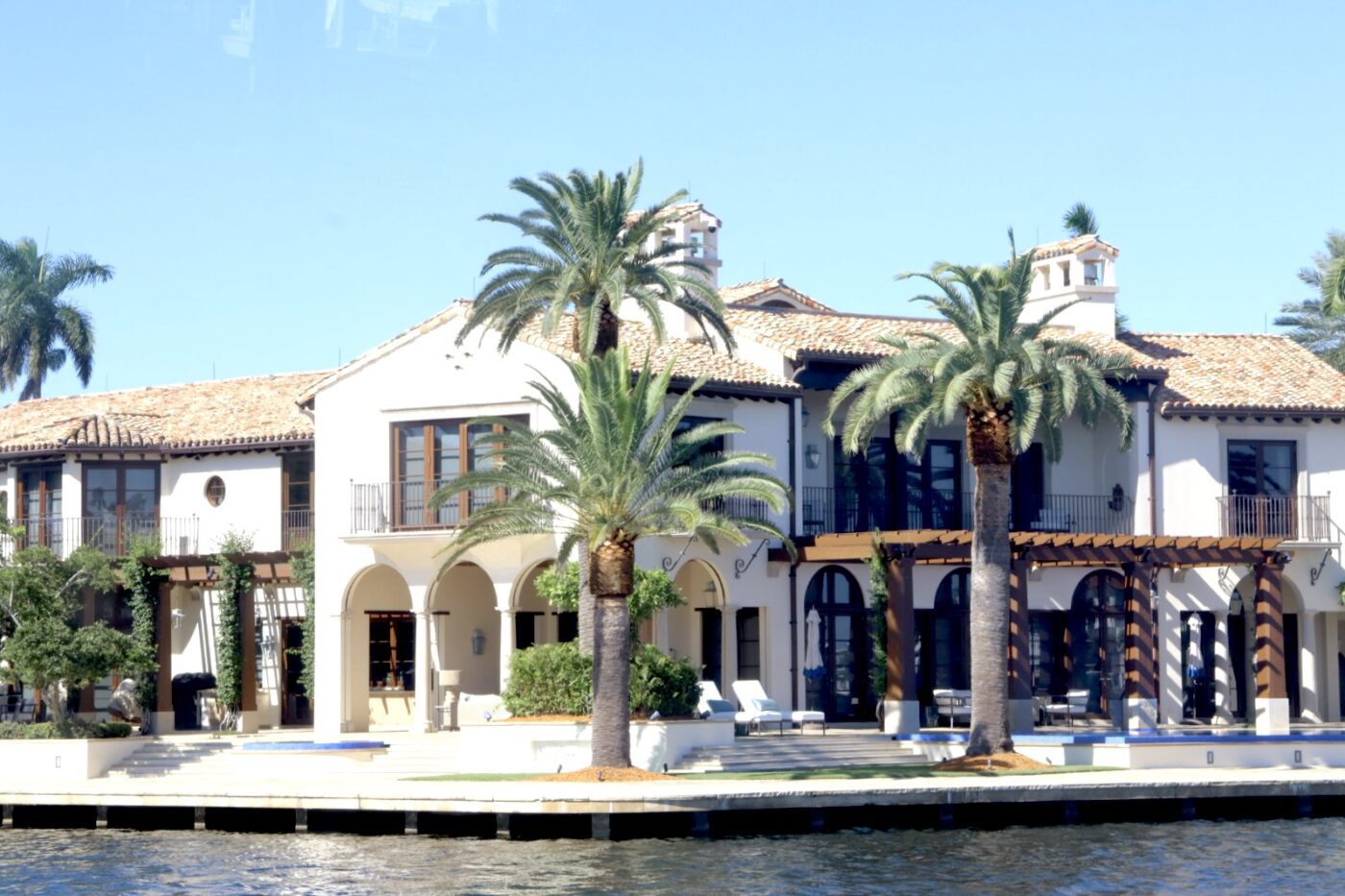 Stylish waterfront villa with palm trees, arched entries, and a tiled roof, showcasing luxury living in a sunny coastal setting.