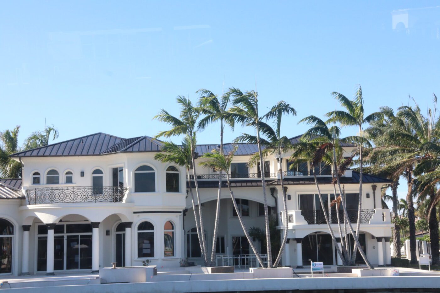 Luxurious waterfront home featuring modern architecture with palm trees, large windows, and a stylish balcony, set against a clear blue sky.