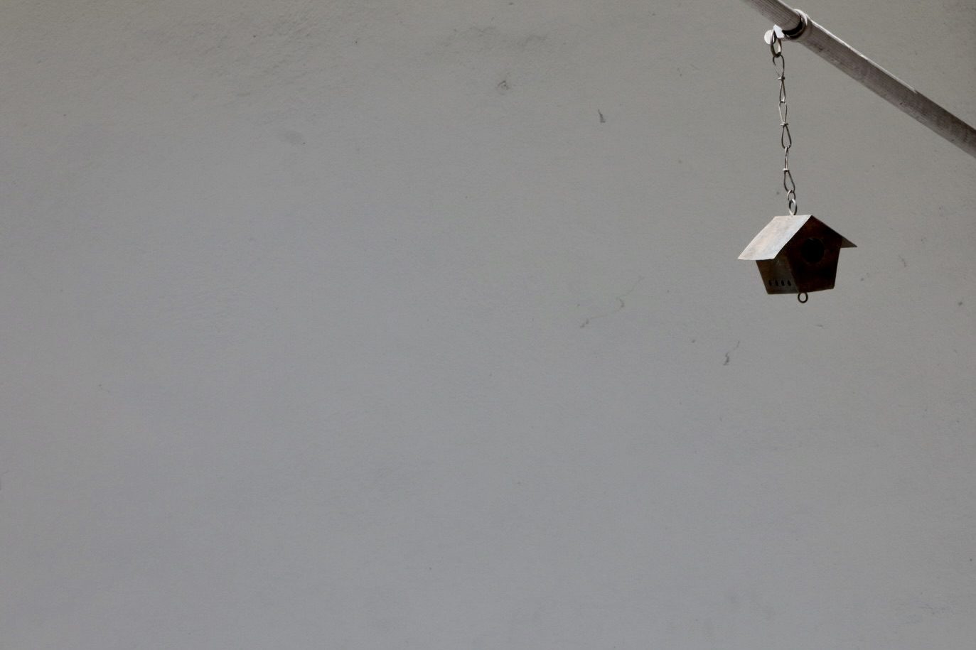 A small metal birdhouse hangs from a chain against a plain gray wall, emphasizing its minimalist design and inviting nature-focused themes.