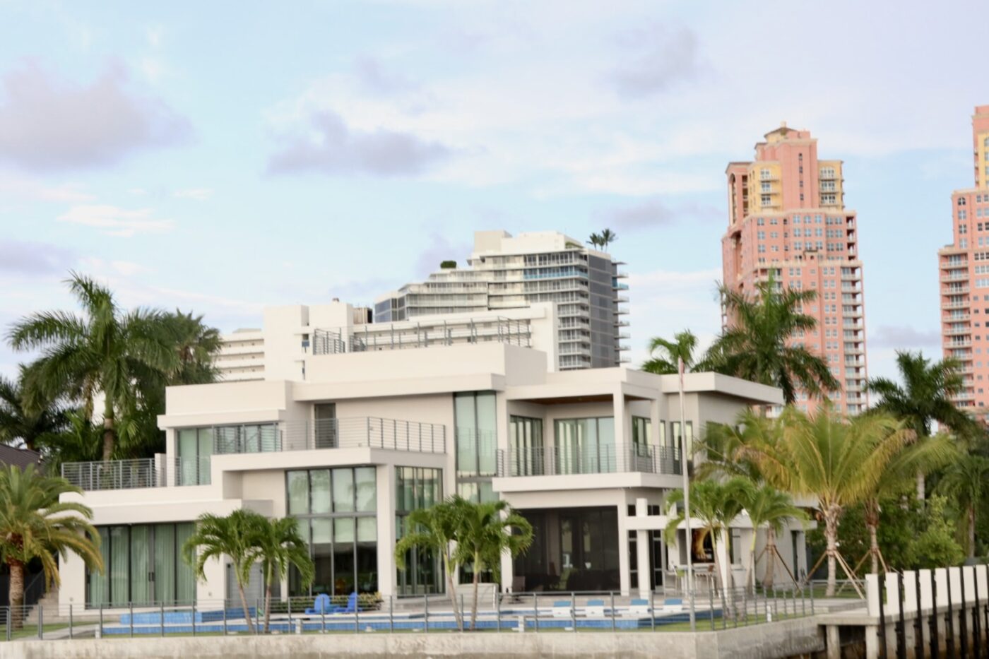 Modern waterfront house featuring large glass windows, palm trees, and a swimming pool, set against a backdrop of urban high-rises.