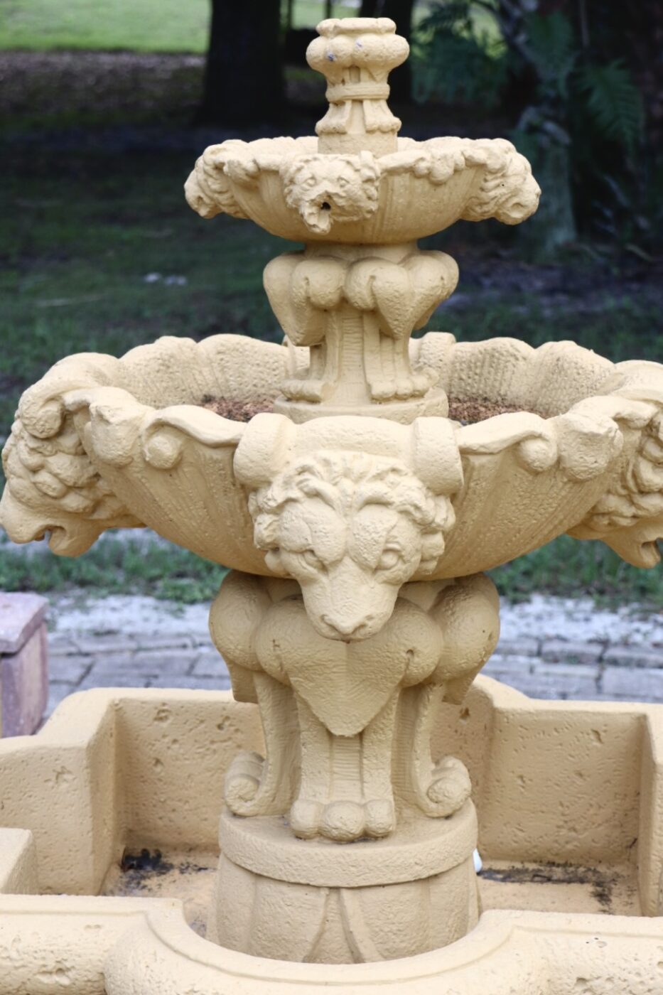 Ornate yellow stone fountain featuring stylized lion heads, set in a garden environment. Ideal for enhancing outdoor aesthetics.