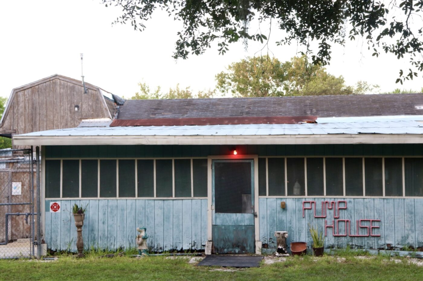 Weathered blue building with "PUMP HOUSE" in neon, featuring a red light above the door, surrounded by greenery and garden decor.